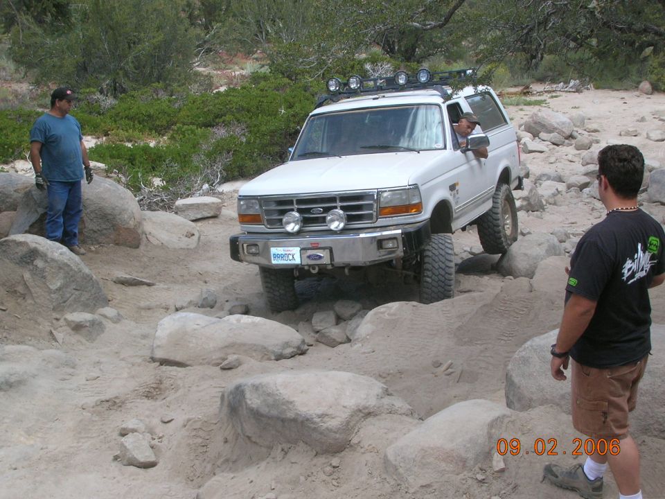 White Ford Bronco navigating rocky terrain. Driver visible. Two men standing nearby