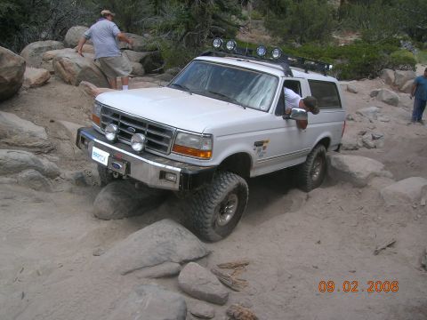 1985 Ford Bronco - A white Ford Bronco is shown off-roading on a sandy, rocky trail with trees in the background.