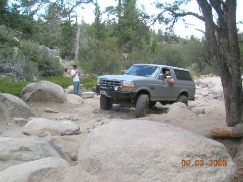 1985 Ford Bronco - A gray SUV tackles a challenging off-road course on a sunny day. The vehicle appears to be from the early to mid-1990s.