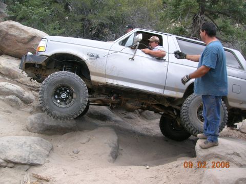 1985 Ford Bronco - A silver Ford Bronco is shown off-roading on a rocky trail with a person guiding it.