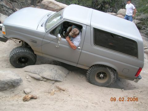 1985 Ford Bronco - A silver 1985 Ford Bronco navigates a rocky, sandy trail with a driver inside and another person observing.