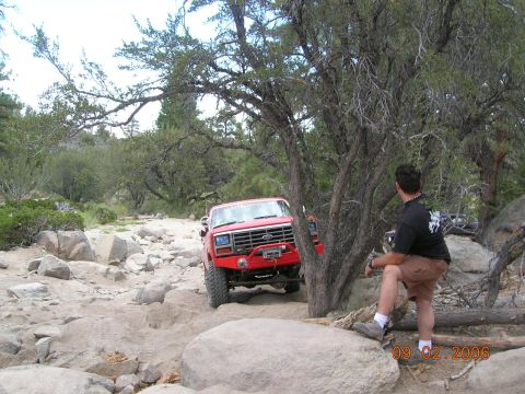 1985 Ford Bronco - A red Ford Bronco is seen off-roading on a rocky path. A man observes from the side.