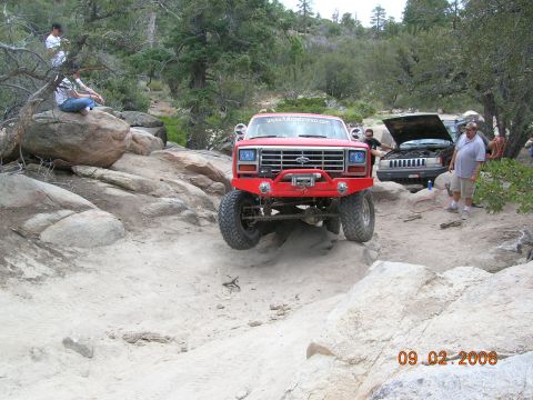 1985 Ford Bronco - A red Ford Bronco navigates a challenging off-road course with other vehicles and people present.