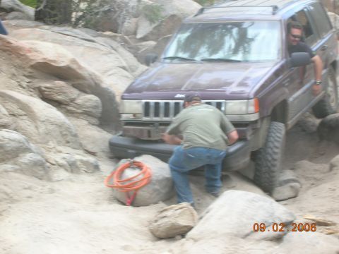 1985 Ford Bronco - A Jeep Grand Cherokee is being driven off-road over rocks and sand, with assistance from a passenger.