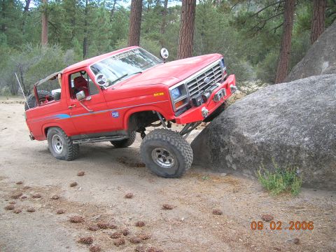 1985 Ford Bronco - Modified red Ford Bronco navigating a rocky trail in a wooded area.