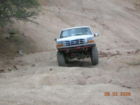 1985 Ford Bronco - White Ford Bronco with lifted suspension driving on a dirt hill. Red aftermarket bumper visible.
