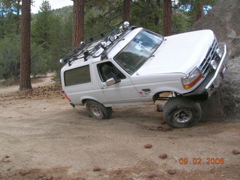 1985 Ford Bronco - White Ford Bronco with roof rack and off-road tires climbing over a large rock on a dirt trail.