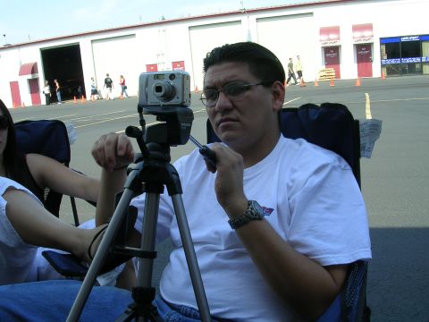1996 Ford Probe - A man adjusts a camera on a tripod while sitting in a chair outdoors, with a building in the background.