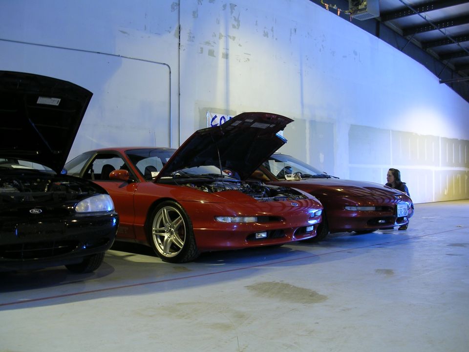 Three Ford Probes parked indoors with hoods open. The front car is black, the middle and rear cars are red. A person stands near the rear car