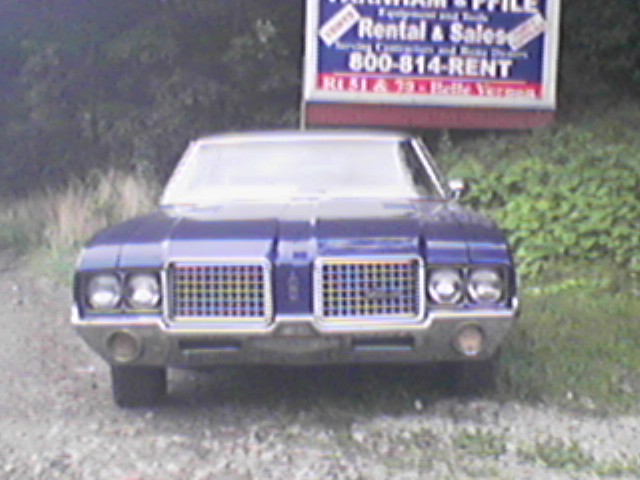 Front view of dark blue vintage Oldsmobile Cutlass with dual headlights and split grille on gravel lot