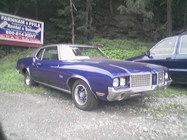 Blue Oldsmobile Cutlass coupe parked on gravel by a rental company sign