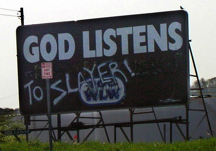 Roadside billboard reading GOD LISTENS with TO SLAYER graffiti spray-painted across it