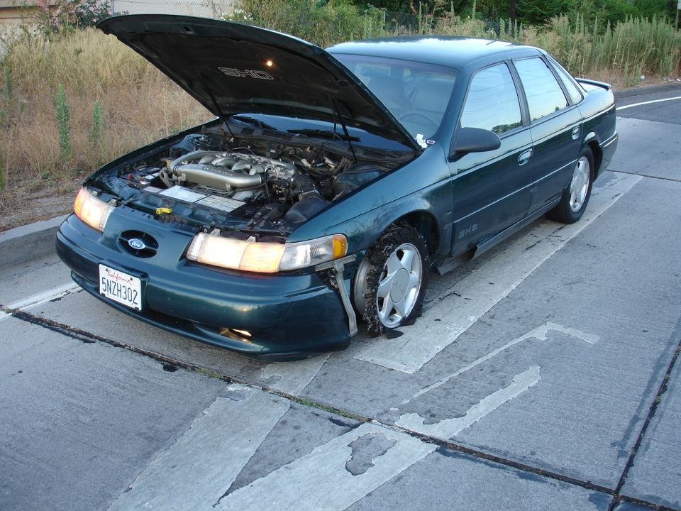 1995 Ford Taurus with hood open, flat front tire, and damaged bumper
