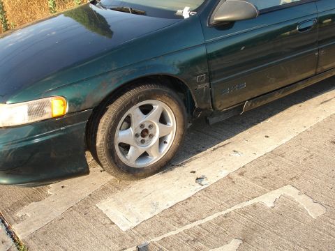 1995 Ford Taurus - A green 1995 Ford Taurus sedan is viewed from the front quarter, revealing damage to its front end.