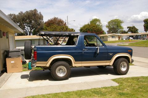1986 Ford Bronco - A blue and tan Ford Bronco with a soft top frame is parked on a driveway next to a house.