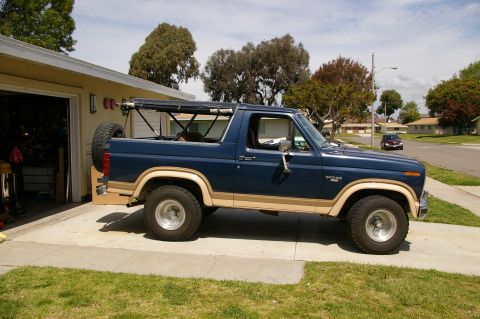 1986 Ford Bronco - This blue Ford Bronco features tan side trim and a visible soft top frame, parked in a driveway.