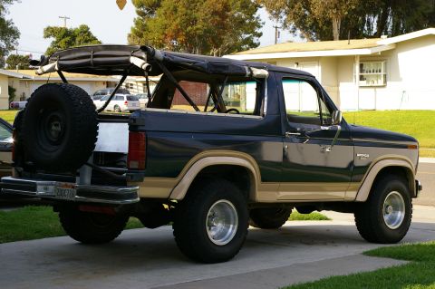 1986 Ford Bronco - This Ford Bronco is parked in a driveway with a house and trees in the background. The soft top is retracted.