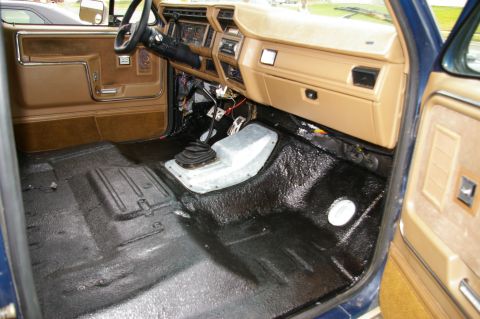 1986 Ford Bronco - Interior passenger-side view of a Ford truck showing a freshly applied black bedliner-style coating on the floor pan, with tan dashboard, d…
