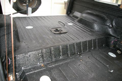 1986 Ford Bronco - Interior of a truck bed or cargo area sprayed in textured black bedliner coating, covering the ribbed floor, the raised step-up section and…