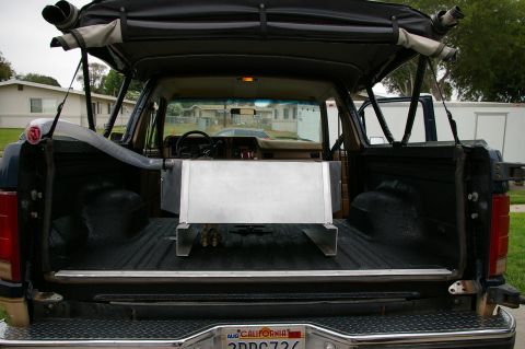 1986 Ford Bronco - Rear view into the open cargo area of a dark blue full-size SUV with the tailgate down, hardtop removed and a soft-top rolled up on the rol…