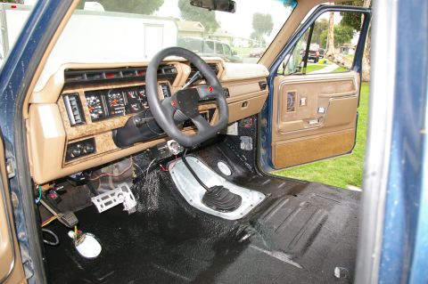 1986 Ford Bronco - Interior view from the passenger side of a Ford Bronco showing the tan dashboard with woodgrain trim, black steering wheel, and a floor-mou…