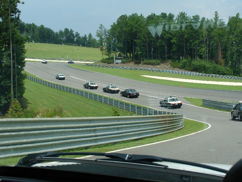 Several police cars driving on a race track with trees and grassy hills in the background