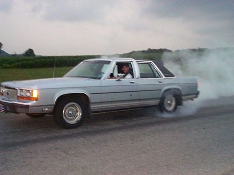 1991 Ford LTD Crown Victoria - A light blue/silver Ford Crown Victoria four-door sedan doing a burnout on a rural road, with a large cloud of tire smoke billowing from th…