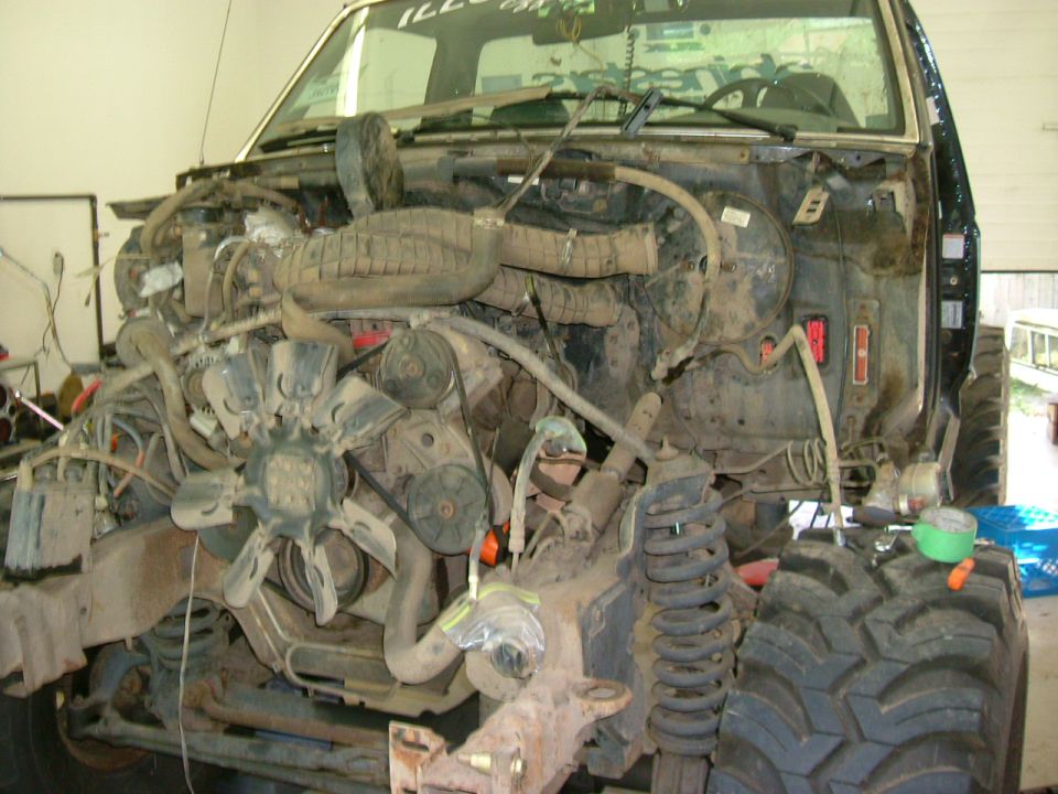 Front of a dark pickup with front clip removed exposing engine, fan, and suspension in a garage