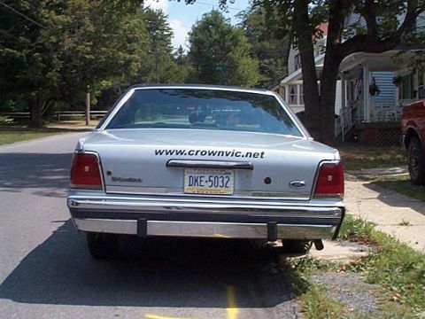1991 Ford LTD Crown Victoria - Rear view of a silver full-size Ford sedan parked at the curb on a tree-lined residential street, with a large 'www.crownvic.net' decal acr…