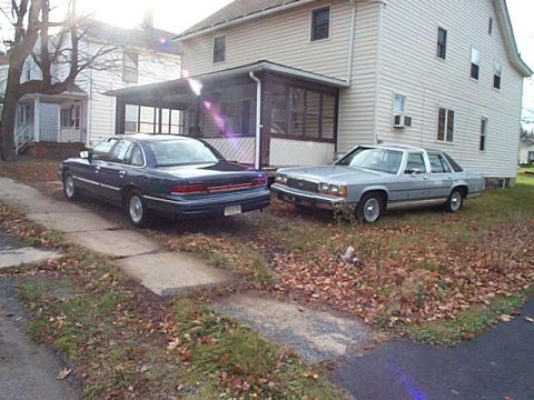 1991 Ford LTD Crown Victoria - Two full-size sedans parked side by side on a driveway beside a white two-story house in autumn: a dark blue Ford sedan seen from the rear…