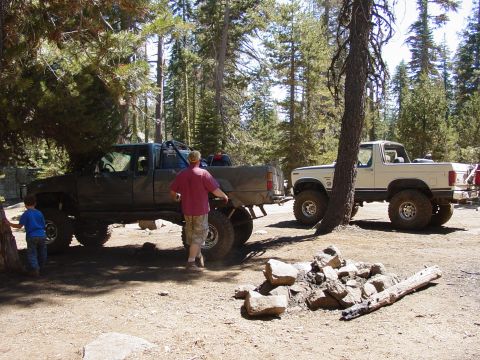1986 Ford Bronco - Two off-road vehicles are parked in a forest clearing, with people nearby.