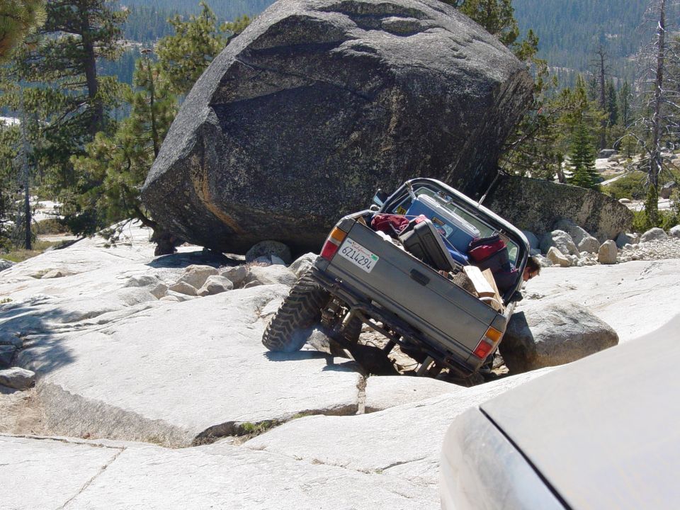 Gray off-road vehicle with a lifted suspension is tilted on rocks, carrying luggage in its bed