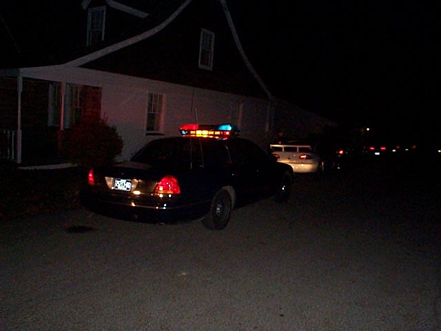 Dark police sedan at night with lit red and blue lightbar parked in front of a house