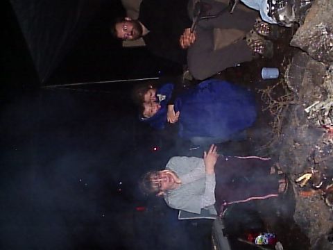 2000 Ford E350 - A group of individuals are seen sitting around a campfire, with smoke drifting through the scene.
