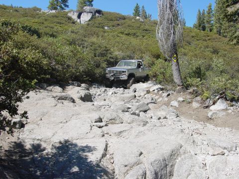 1986 Ford Bronco - A silver pickup truck navigates a challenging rocky terrain on a sunny day.