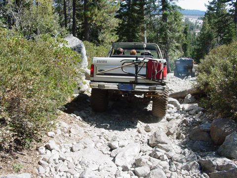 1986 Ford Bronco - Two vehicles navigate a challenging, rocky off-road trail through a wooded mountain area.