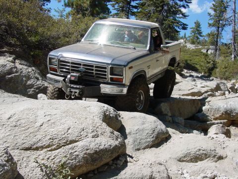 1986 Ford Bronco - This modified pickup truck navigates a rocky off-road path under a clear blue sky.