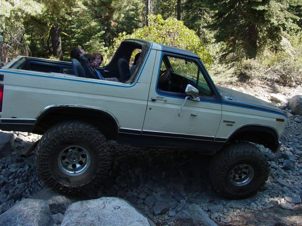 White Ford Bronco with blue trim, lifted with large tires, driving over rocks. Passengers in the back