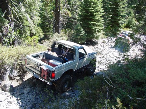 1986 Ford Bronco - This modified SUV navigates a challenging, rocky path in a wooded area, with another vehicle in the distance.