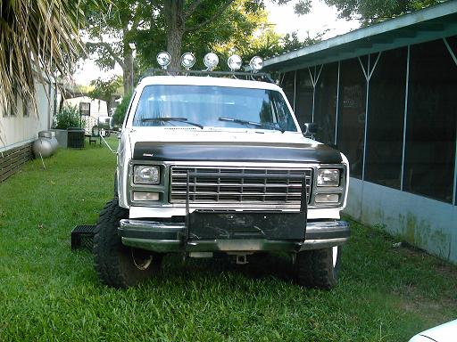 Front view of a lifted white pickup truck with a black hood and off-road lights on the roof