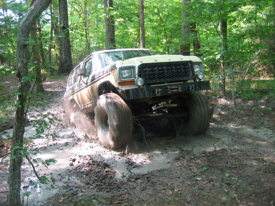 Modified Ford Bronco or F-Series truck with oversized tires driving through mud and water in a wooded area