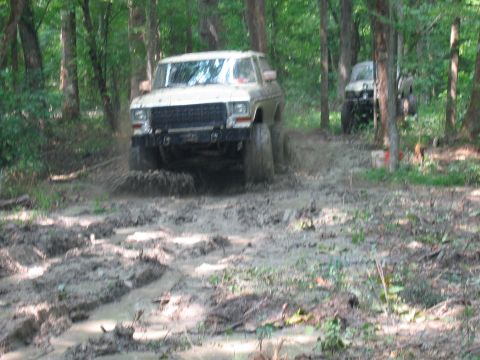 1992 Ford Mustang - A white 1979 Ford Bronco navigates a muddy trail in a wooded area, with another vehicle visible in the background.