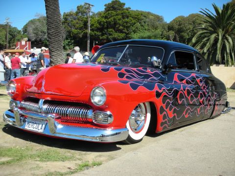 1998 Ford Crown Victoria - chopped Mercury lead-sled custom in black with red-and-blue flames sits low on whitewall tires at an outdoor car show under palms.