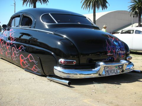 1998 Ford Crown Victoria - Rear three-quarter of a black chopped Mercury custom with flame graphics, lakes pipe, and a California MERCULA license plate.