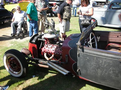 1998 Ford Crown Victoria - rat-rod roadster pickup with an exposed red engine, headers, and whitewall tires sits on grass as onlookers gather at a car show.