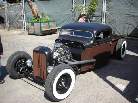 1998 Ford Crown Victoria - chopped, lowered black rat-rod pickup with a chromed V8 and tall whitewall tires sits on pavement against a green tarp backdrop.