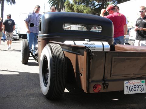 1998 Ford Crown Victoria - Rear three-quarter of a low black rat-rod pickup with a wide whitewall tire and a California plate reading 7X65931, with onlookers nearby.
