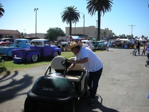 1998 Ford Crown Victoria - man in a straw hat leans over a green Club Car golf cart among rows of vintage pickups and palms at a sunny outdoor car show.