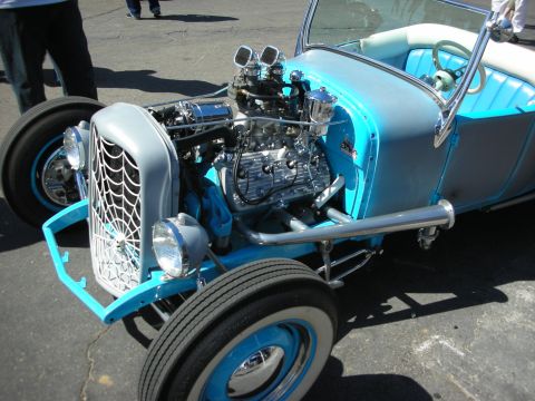 1998 Ford Crown Victoria - light-blue open roadster hot rod shows off an exposed supercharged V8 and a spiderweb grille, parked on pavement at a show.