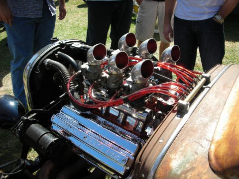 1998 Ford Crown Victoria - polished V8 engine fitted with six chrome velocity stacks and red ignition wires sits in an open hot rod engine bay as spectators stand by.